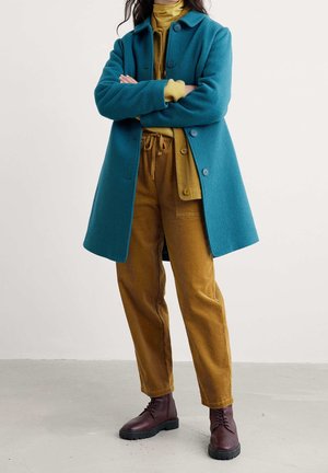 Personne portant un manteau bleu sarcelle sur une tenue superposée jaune moutarde, un pantalon en velours côtelé et des bottines lacées marron foncé, debout les bras croisés.
