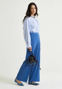Pantalon large bleu en tissu texturé, associé à un chemisier rayé bleu et blanc. Les accessoires comprennent un petit sac noir perlé et des chaussures à motif.