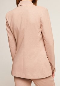 Bledě růžový blazer s lesklou texturou, strukturovaným designem, klopami a zapínáním na knoflíky, který ukazuje přizpůsobený střih a čisté linie.