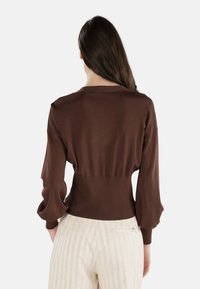 Brązowy sweter z długim rękawem, dopasowany w talii z prążkowanym wykończeniem oraz falbanami przy mankietach, w połączeniu z jasnymi spodniami w paski. Gładka tekstura materiału.