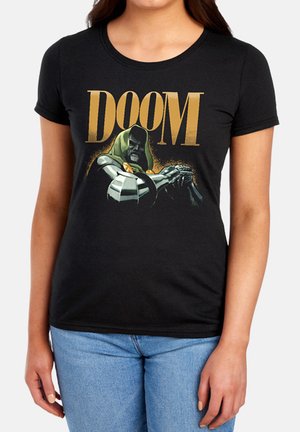 Frau, die ein schwarzes T-Shirt mit goldenem "DOOM" Text und einer Illustration einer Figur in grüner Kapuze und Metallrüstung trägt, die die Arme verschränkt.