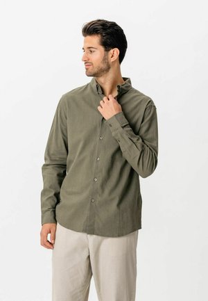 Mężczyzna poprawiający kołnierz oliwkowozielonej koszuli na długi rękaw z guzikami, ubrany w jasnobeżowe spodnie, stojący na białym tle.