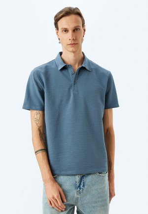 Kurzarm-Poloshirt aus strukturiertem, hellblauem Stoff. Mit klassischem Kragen und einer drei-knöpfigen Knopfleiste. Getragen mit verblasstem Denim.