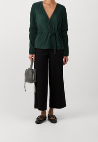 Donna che indossa un top avvolgente plissettato verde scuro con fiocco laterale, pantaloni neri a gamba larga, scarpe basse nere con dettagli in fibbia, mentre tiene una borsa in argento texturizzato.
