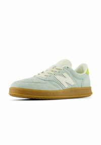 Jasnozielone zamszowe sneakersy z białym logo New Balance, kremowymi sznurowadłami i gumową podeszwą. Posiadają perforacje na palcach oraz neonowy akcent na pięcie.