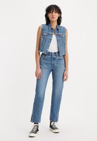Denim vest with front button closure and two chest pockets, paired with high-waisted straight-leg jeans featuring frayed hems and side embellishments.