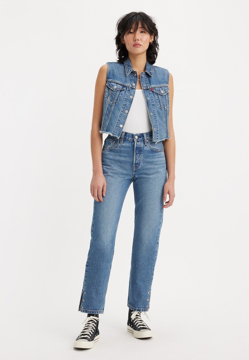 Denim vest with front button closure and two chest pockets, paired with high-waisted straight-leg jeans featuring frayed hems and side embellishments.