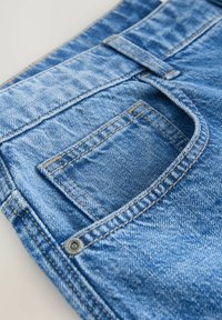 Modre denim hlače imajo vidno zadnjo žep, kontrastno rumeno šivanje in kovinsko zakovicno okrasje ob robu žepa.