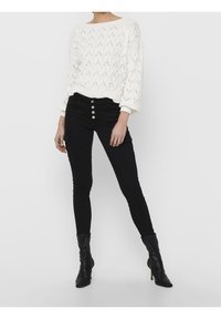 Witte gebreide trui met een gestructureerd, open geweven patroon, gecombineerd met zwarte skinny jeans met een hoge taille en zilveren knoopdetail. Zwarte enkellaarzen.