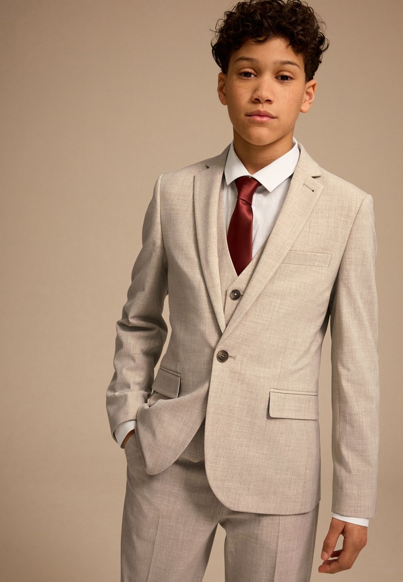 Costume gris clair à finition texturée, avec une veste à deux boutons, des revers à crans, et une cravate bordeaux. Chemise blanche en dessous.