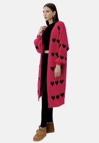 Lange roze gebreide cardigan met zwarte hartpatronen, open voorkant en brede mouwen. Combineer met zwarte leggings en tan enkellaarsjes.