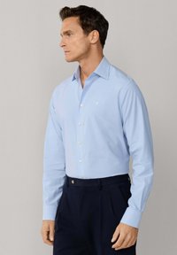 Muž v světle modré košili s dlouhým rukávem na knoflíky a tmavě modrých kalhotách stojí bokem na jednobarevném šedém pozadí.