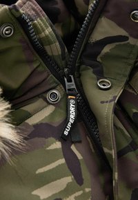 Kamerová bunda s predným zipsom so značkovaním "SUPERDRY", kožušinovým lemovaním goliera a kovovými cvokmi. Textúrovaná látka v odtieňoch zelenej a hnedej.