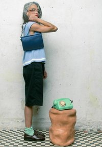 Top sin mangas azul acanalado con ribete blanco, pantalones cortos negros hasta la rodilla, y los accesorios incluyen un bolso azul y zapatos marrones. Teléfono rotatorio verde sobre un taburete.