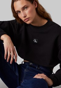 Felpa nera corta con maniche lunghe, colletto a costine, con una piccola toppa del logo Calvin Klein. Indossata con jeans blu scuro.