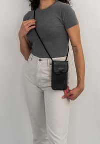 Borsa a tracolla nera in materiale sintetico con una patta testurizzata simile a un coccodrillo, caratterizzata da un logo dorato e una lunga tracolla. Indossata all'anca.