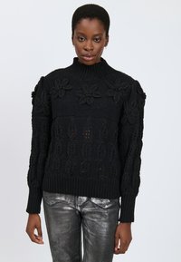 Maglione nero a maglia con collo alla coreana, con dettagli floreali e motivi testurizzati. Polsini e orlo a coste. Indossato sopra pantaloni metallici argento.