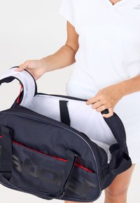 Person i hvid kjole, der åbner en tom sort og rød sportstaske med stort "Babolat" branding på frontlommen.