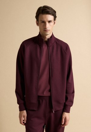 Joven vestido con un chándal granate con chaqueta con cremallera, de pie frente a un fondo beige liso.