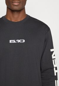 Črna majica s dolgimi rokavi iz bombaža, z majhnim belim vezenim logotipom "5.10" na prsih in belim napisom ob desnem rokavu.