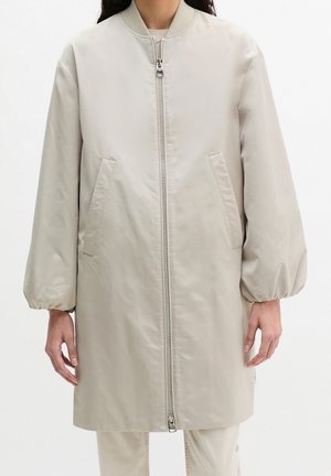 Femme portant une longue veste bomber beige avec poches avant, col côtelé, fermeture éclair frontale et poignets élastiques aux manches longues.