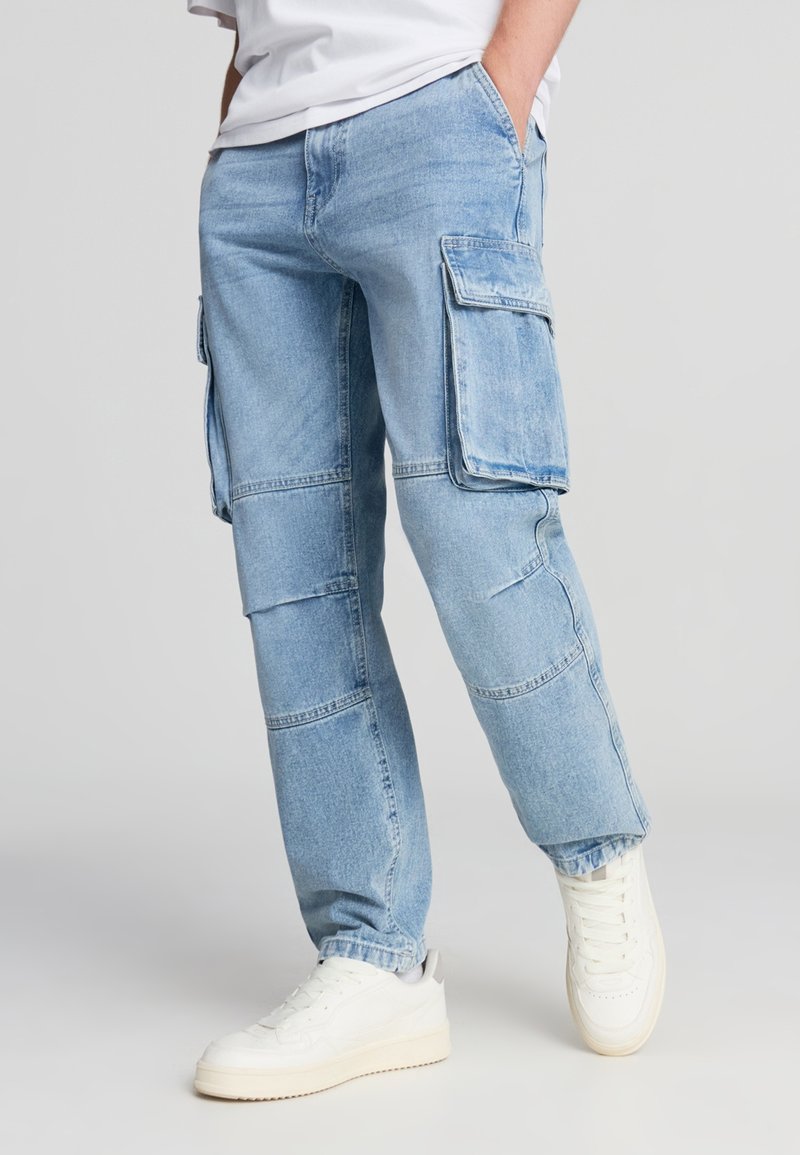 Pantaloni cargo azzurri chiari in denim, caratterizzati da numerose tasche grandi e una vestibilità comoda, abbinati a sneaker bianche.