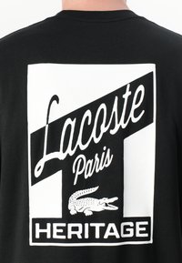 Svart t-skjorte med en stor hvit grafikk på baksiden med teksten "Lacoste Paris" og "HERITAGE", sammen med en krokodilleillustrasjon.