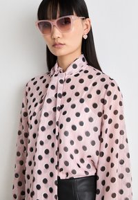 Lys rosa gjennomsiktig bluse med svarte prikker, med høy krage og lange ermer. Modellen har på seg rosa oversized solbriller og hjerteformede øreringer.