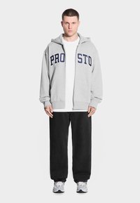 Osoba stoi przodem, ubrana w szary polar na zamek z napisem "PRO", białą koszulkę, czarne spodnie i tenisówki, na białym tle.