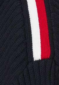 Námornícky pletený sveter s ribovanou texturou, s vertikálními pruhy v červené, bílé a červené barvě umístěnými podél ramenního švu.