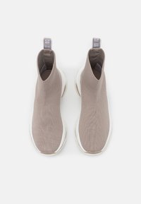 Botas de tobillo tricotadas en color beige con parte superior elástica, suela blanca y una pestaña en bucle gris en la parte posterior. Textura suave y diseño ligero.