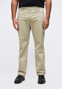 Homme portant un pantalon beige droit et des chaussures noires, debout contre un fond clair uni, les mains détendues le long du corps.