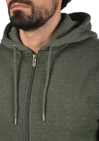 Groene rits-hoodie gemaakt van zachte stof, met een trekkoordcapuchon en een zilveren metalen rits met een gestructureerde trekker.
