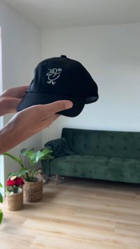 Black cap held in a hand, featuring a white embroidered bird design on the front. Soft fabric with a curved brim and adjustable strap.