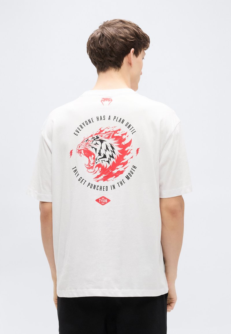 Λευκό T-shirt με κοντά μανίκια, με ζωντανό γραφικό ενός βρυχώμενου τίγρη σε κόκκινο και μαύρο, με κείμενο και λογότυπο στην πλάτη.