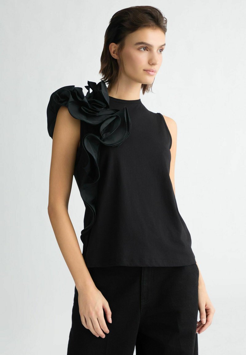 Top nero senza maniche con un grande accentuato arricciato sulla spalla sinistra, realizzato in un tessuto liscio con una vestibilità comoda. Indossato con pantaloni scuri.