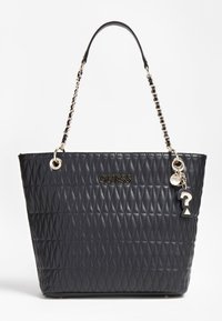 Sac de transport matelassé noir avec chaîne et sangle en cuir, logo GUESS doré, et charmes pendants incluant un point d'interrogation et une étiquette ronde.