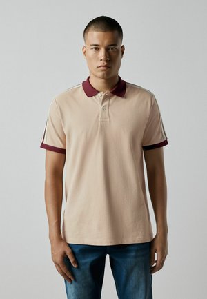 Ung mand med kort hår iført en beige poloshirt med vinrød krave og ærmekanter, stående mod en ensfarvet lys baggrund.