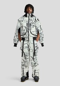 Férfi, aki fehér síkabátot és nadrágot visel fekete, absztrakt graffitistílusú mintával és fekete síbakancsokkal, zsebre tett kézzel áll.