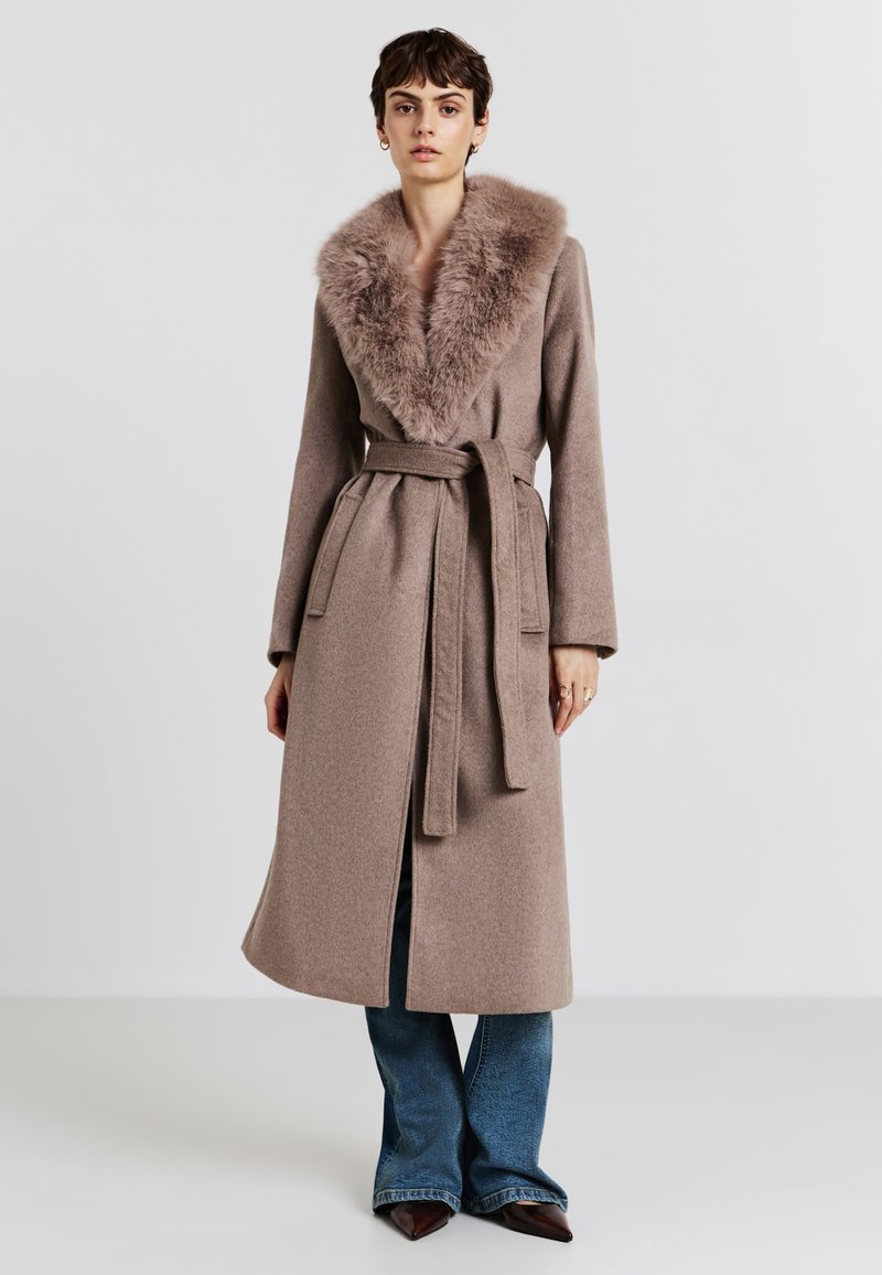 Cappotto di lana marrone con colletto in pelliccia sintetica, vita con cintura, maniche lunghe e tasche laterali, indossato sopra jeans a zampa d'elefante.