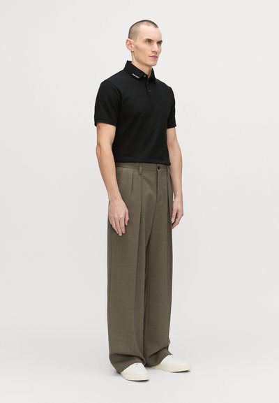 Black short-sleeve polo shirt with collar branding, paired with wide-legged light brown trousers and white sneakers. Plain background.