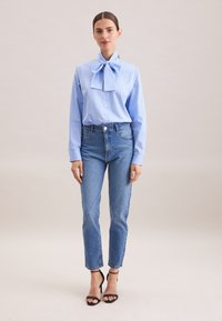 Hellbluse mit Streifen und einer großen Schleife, kombiniert mit hochgeschnittenen, gerade geschnittenen blauen Jeans und schwarzen, hochhackigen Sandalen. Einfaches, sauberes Design.