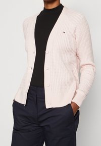 Heller rosa Cardigan mit gerippter Textur, ausgestattet mit einer knöpfbaren Vorderseite, zwei Taschen und einem kleinen Logodetail auf der Brust.