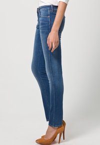 Mörkblå skinny jeans med medelhög midja, tillverkade av slitstarkt denimmaterial med subtila sömnadsdetaljer. Bärs med spetiga beige klackar.