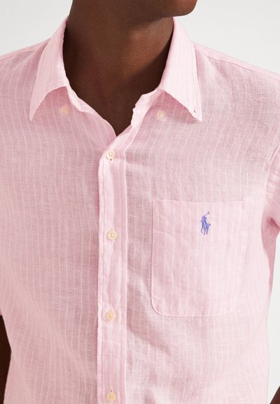Detailný záber na osobu nosiacu svetloružovú ľanovú košeľu s vertikálnymi pruhmi, náprsným vreckom a malým modrým vyšívaným logom.