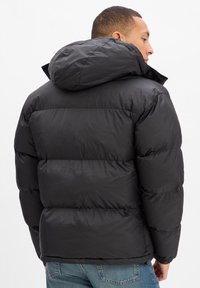 Schwarze Steppjacke mit Kapuze, die ein gesteppte Design und eine glatte, glänzende Textur aufweist. Die Rückseite zeigt elastische Bündchen und eine lockere Passform.