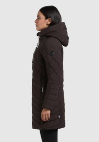 Brun, quiltet parka med hætte, der har et diagonalt mønster, lynlåslommer og en slank silhuet. Sorte accenter og et logo-patch.