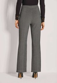 Frau, die hochtaillierte schwarz-weiß gemusterte weit geschnittene Hose und schwarze Absatzstiefeletten trägt, vor einem schlichten Hintergrund steht.