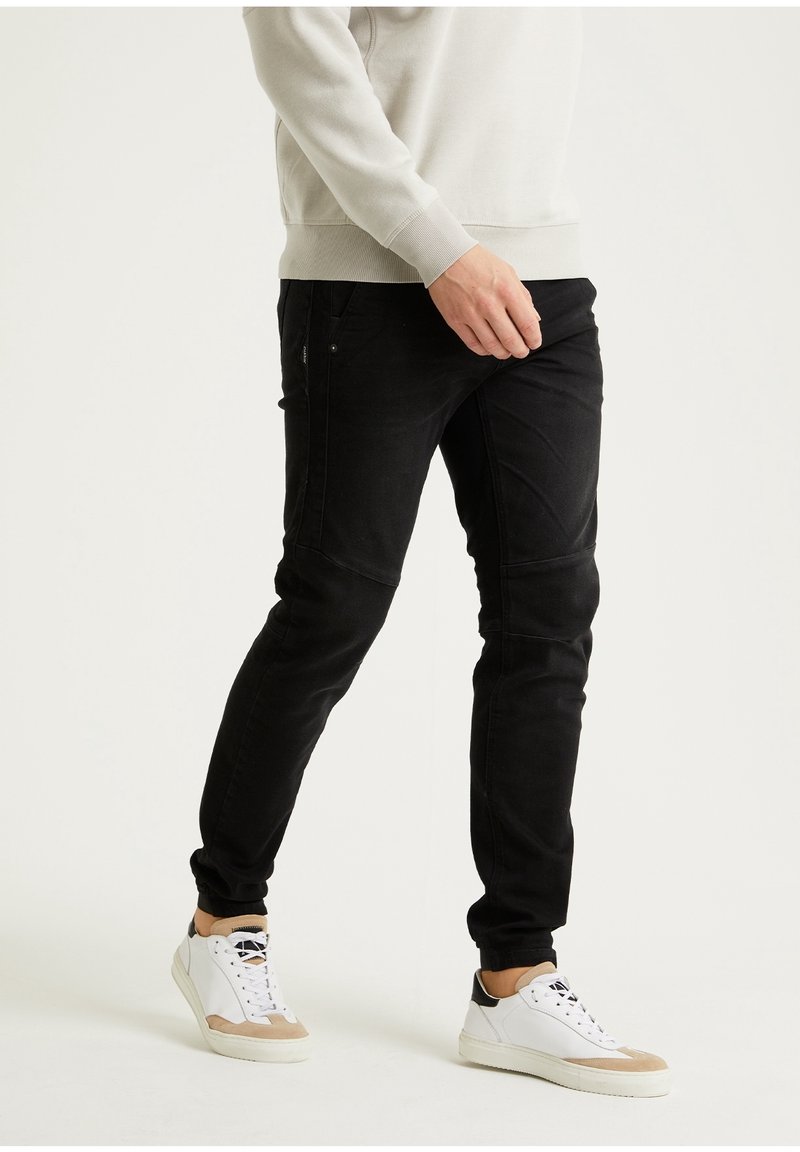 Pantalones de mezclilla negros con un ajuste cónico, que presentan detalles cosidos en las rodillas y bolsillos laterales, combinados con zapatillas blancas y una sudadera gris claro.