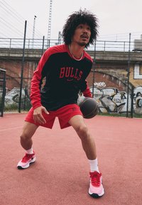 Red and black athletic outfit with "BULLS" text, featuring long sleeves and shorts. White socks and red sneakers, holding a basketball.